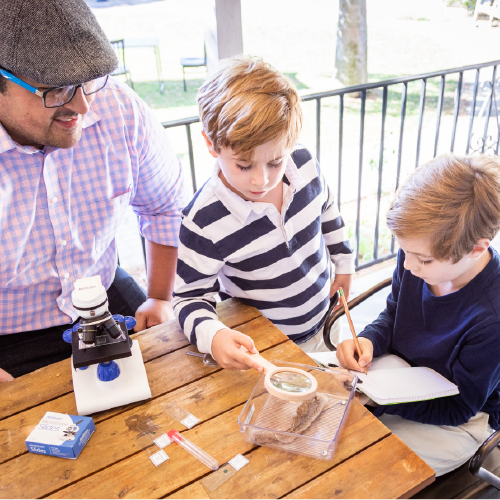 Male tutor sitting with grammar school boys at a table using magnifying glasses to examine plants